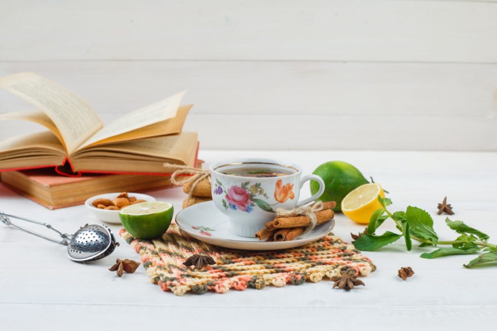 high angle view a cup of tea with cinnamon and lemon on square placemat with limes,a bowl of almonds,tea strainer and books on white background. horizontal free space for your text
